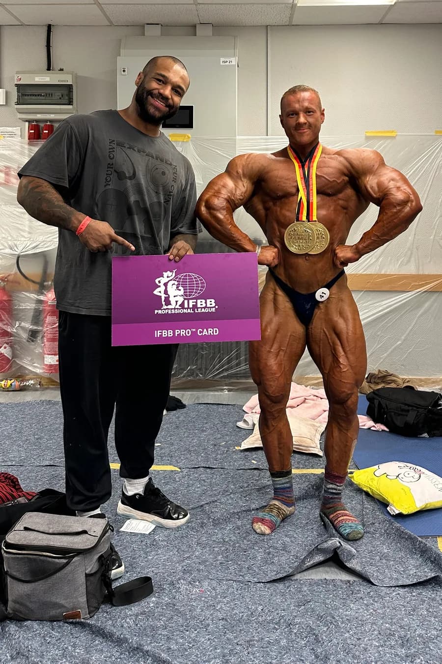 Dwayne hält das IFBB-Pro-Card-Schild, daneben Dennis mit zwei Medaillen